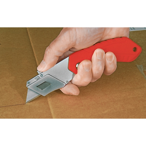 A knife is cutting cardboard. The handle is red, the metal is silver. The hand is holding the knife.
