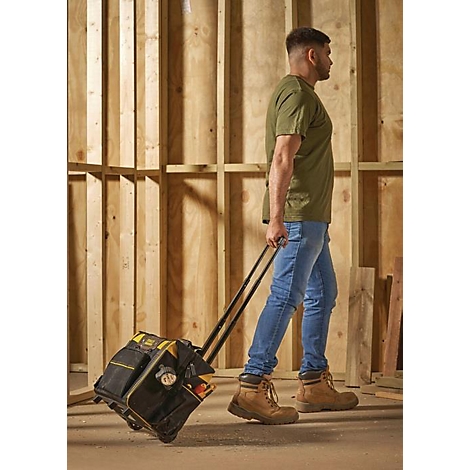 Ein Mann geht mit einer schwarzen Werkzeugtasche auf Rädern. Er trägt Jeans und ein grünes T-Shirt. Hinter ihm ist eine Holzkonstruktion.