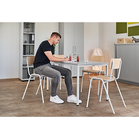 Een man zit aan een tafel te schrijven. Er staan drankjes op tafel. Stoelen, kast, lamp op de achtergrond.