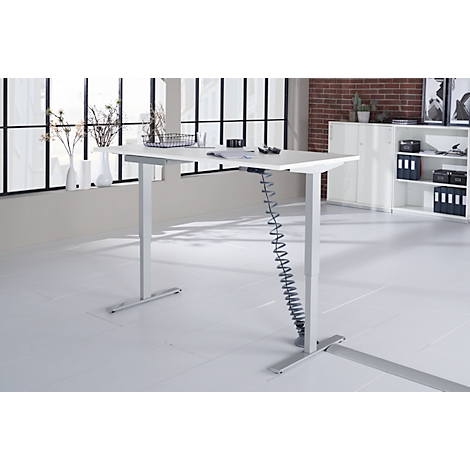 Height-adjustable desk. White tabletop, gray legs, behind it windows. Brick wall and cabinet in the background.