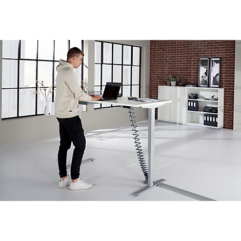A man stands at a height-adjustable desk, working on a laptop, light clothing. A shelf and a window in the background.