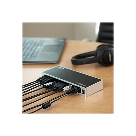 Ein schwarzer USB-C-Hub mit mehreren AnschlÃ¼ssen, an dem Kabel angeschlossen sind, liegt auf einem Holztisch neben einem Laptop und KopfhÃ¶rern.