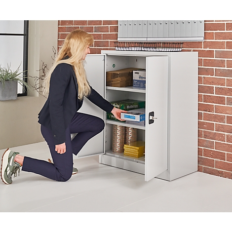 A woman kneels in front of a white cabinet, opens it, and reaches for an item.