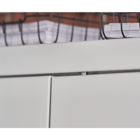 Close-up of white cabinet doors, a narrow silver metal trim and a wire basket with papers in the background.