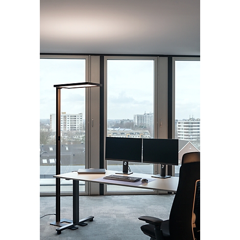 Büro mit Schreibtisch, Monitoren und moderner Stehlampe; Blick durch Fenster auf Stadt.