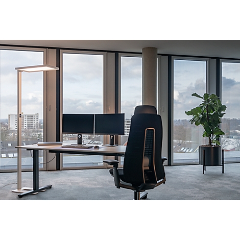 Büro mit Fensterfront, Schreibtisch, Monitoren, Stehleuchte und Bürostuhl mit Blick auf eine Stadt.
