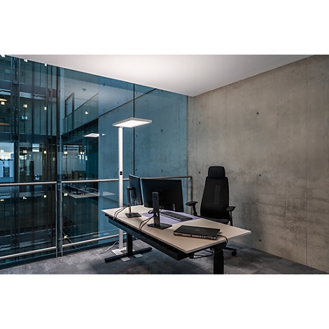 Büro mit Schreibtisch, Monitoren, Stuhl und Stehleuchte. Graue Betonwand und Blick auf eine moderne Glasfassade.