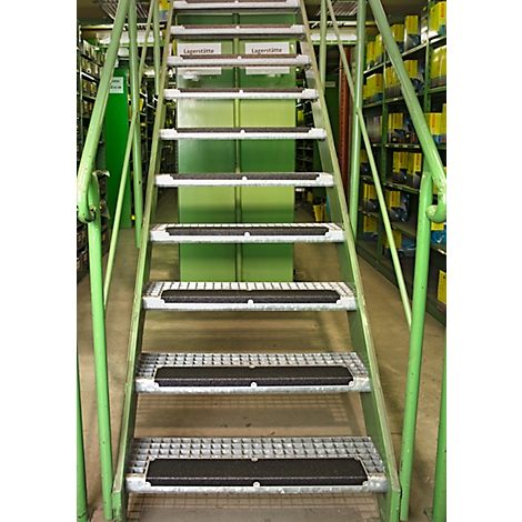 Metal staircase in a warehouse, with green railings and black anti-slip strips on the steps. Shelves in the background.