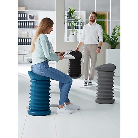 A woman sits on a blue ergonomic stool while a man stands in an office. Two other stools are visible.