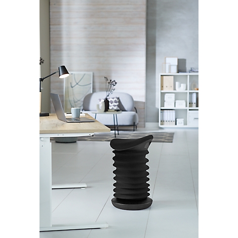 Black standing stool in a minimalist home office. Desk, couch, and shelves are visible in the background.