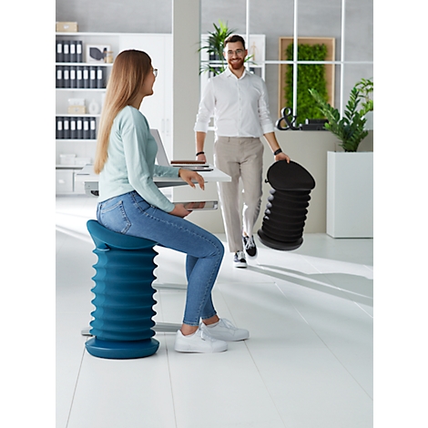 A woman is sitting on a blue stool at a desk, a man is carrying a black stool.
