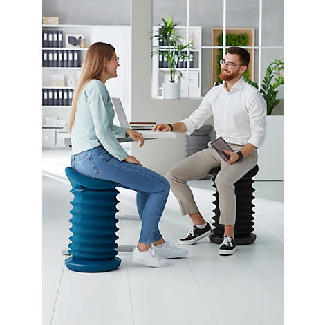 Two people sit on ergonomic stools in an office. They are talking, a woman is smiling.