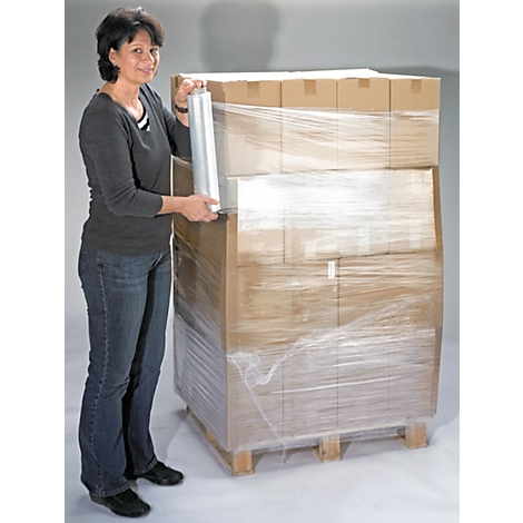 Woman wrapping a pallet of cardboard boxes with plastic wrap. She stands left of the pallet and smiles at the camera.