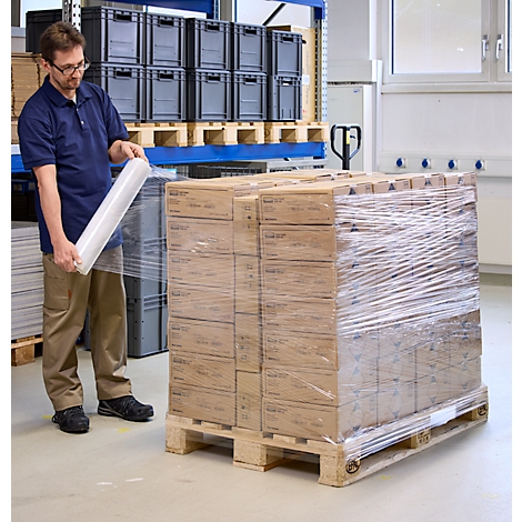 Man wraps a pallet of boxes in foil. He wears glasses, a blue polo shirt and beige trousers.