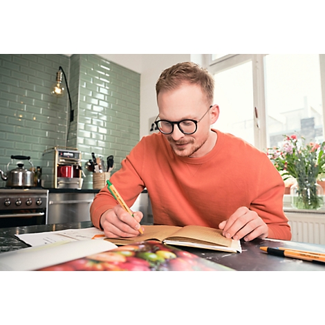 Un homme avec des lunettes et un pull orange est assis en train d'écrire dans un cahier. Environnement de cuisine.