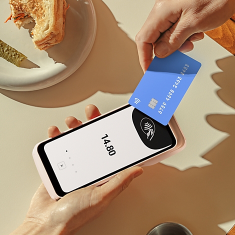 Hände halten ein Smartphone mit 14,80 Euro Anzeige und eine blaue Karte für kontaktloses Bezahlen. Im Hintergrund: Essen.