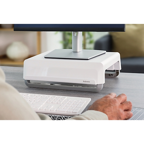 Un support de moniteur blanc est posé sur un bureau gris. Une main repose à côté d'un clavier blanc. Le moniteur est noir.