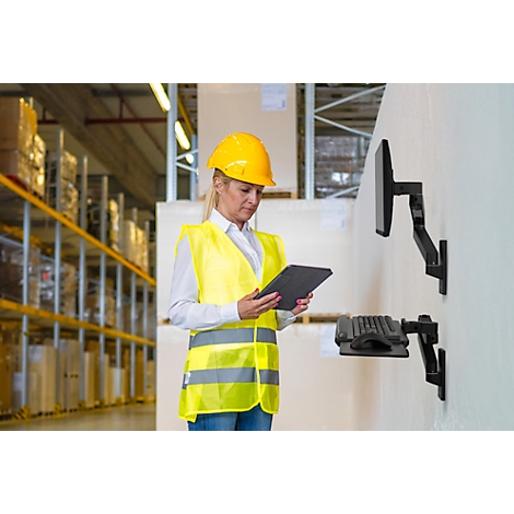 Femme dans un entrepôt portant un casque et un gilet jaunes, tenant une tablette. Étagères et écran en arrière-plan.