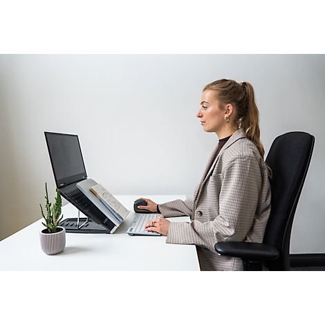 Une femme est assise à un bureau avec un ordinateur portable, une souris et un clavier. Elle porte un blazer, ses cheveux sont attachés en queue de cheval.