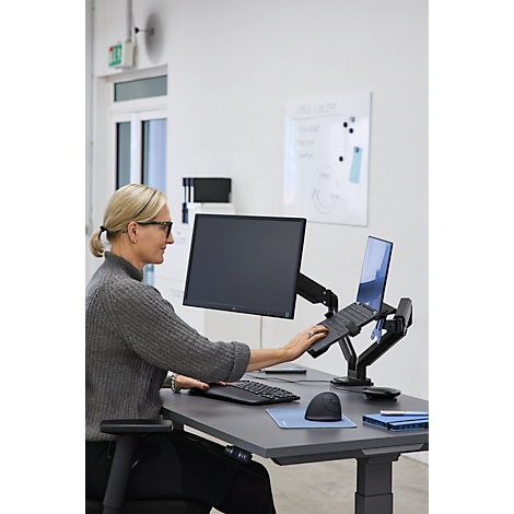 Une femme travaille à un bureau réglable en hauteur avec un moniteur, un ordinateur portable, un clavier et une souris. Elle utilise l'ordinateur portable.
