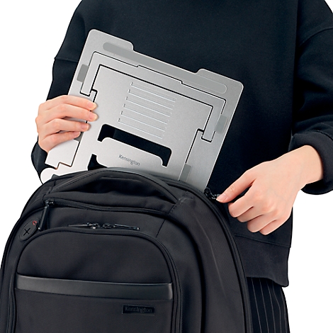 Une personne range un support d'ordinateur portable argenté dans un sac à dos noir. Mains saisissant le support et la fermeture éclair.