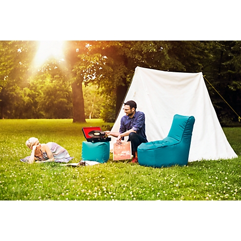 Two people camping: man at record player, woman lying on blanket. Green space, tent, armchair, sunny ambience.