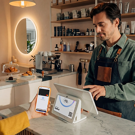 Un barista utilise un terminal de paiement lumineux pendant qu'un client paie sans contact avec un smartphone.
