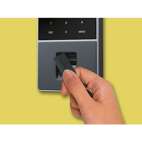 Une main tient un porte-clés devant un lecteur de carte gris fixé à un mur jaune.
