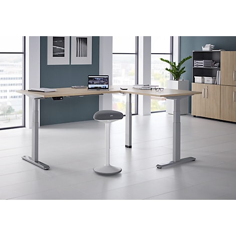 Un bureau d'angle réglable en hauteur avec un ordinateur portable, des lunettes et un tabouret dans un bureau moderne, environnement lumineux.