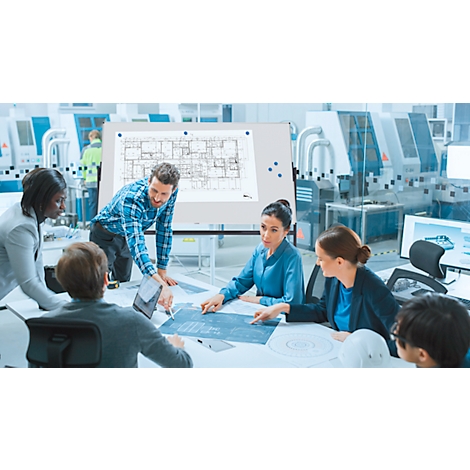 Des ingénieurs examinent des plans à une table. Des croquis et une tablette sont sur la table. Des machines et un plan d'étage en arrière-plan.