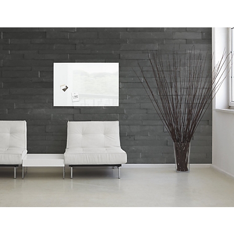 Tableau magnétique en verre blanc sur un mur de pierre sombre, avec deux canapés blancs en dessous. À droite, un vase de fleurs séchées.