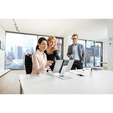 Three people looking at a tablet on a desk in the office. One woman is sitting, two are standing next to her.