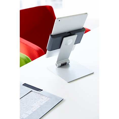A tablet holder with tablet stands on a white table. In the background you can see a red armchair and a document.