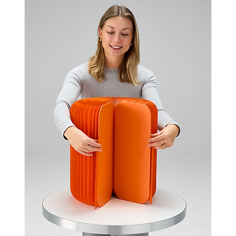 Une femme tient dans ses mains un tabouret orange en forme d'accordéon, sur fond neutre.