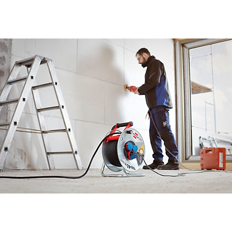 Un homme travaille sur un mur avec une ponceuse. Une bobine de câble et une échelle sont dans l'image.