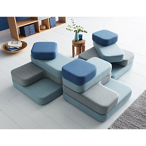 Modular sofa system in blue and grey. Multiple elements form a seating group on a light floor. Shelf and decoration in the background.