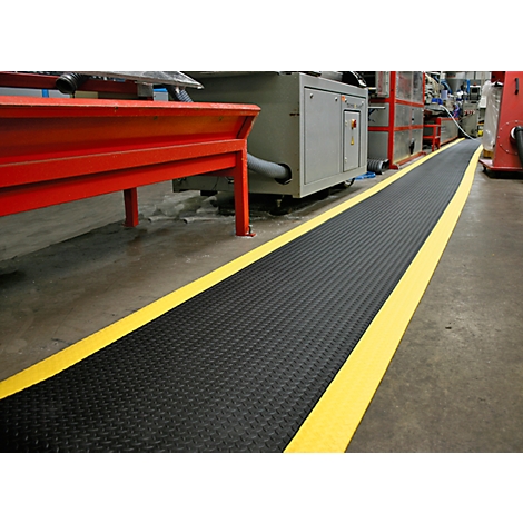 Tapis gaufré noir avec marquages jaunes dans une usine, machines en arrière-plan.