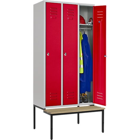 Un armario blanco y rojo con la puerta abierta, ropa y herramientas cuelgan en el interior. Un banco debajo.