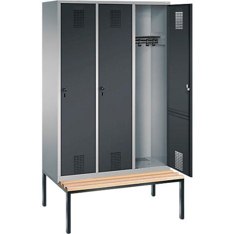 Taquillas metálicas grises, una puerta abierta, con banco de madera delante. Los armarios tienen orificios de ventilación y ofrecen espacio para colgar ropa.
