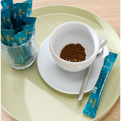 Weiße Kaffeetasse mit Instantkaffeepulver auf einem hellgrünen Tablett, daneben Tchibo Kaffeesticks im Glas.