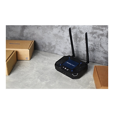 Schwarzer Router mit zwei Antennen. Daneben sind zwei braune Kartons zu sehen. Der Router steht auf einer grauen OberflÃ¤che.