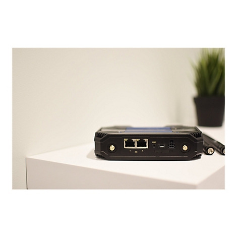 Schwarzer Router mit AnschlÃ¼ssen auf einer weiÃŸen FlÃ¤che. Im Hintergrund Topf mit grÃ¼ner Pflanze und Stift.