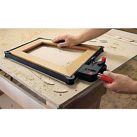 Des mains travaillent sur un cadre en bois avec un connecteur d'angle noir et un outil rouge sur un établi. Des morceaux de bois sont posés à côté.