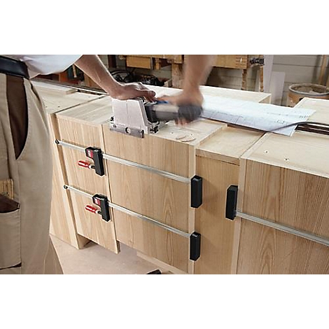 Un charpentier travaille le bois avec une défonceuse sur un meuble en bois. Des pinces maintiennent les pièces ensemble.