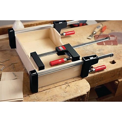 Une boîte en bois est collée avec des pinces. Poignées rouges, supports noirs sur une surface en bois clair.