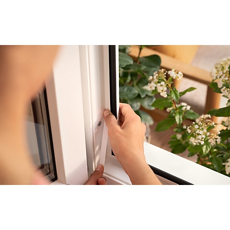 Nahaufnahme einer Person, die eine Dichtung in einem weißen Fensterrahmen anbringt. Im Hintergrund grüne Pflanzen und ein Holztisch.