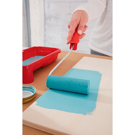 Person painting a wooden board with turquoise paint. Red paint tray on the left.