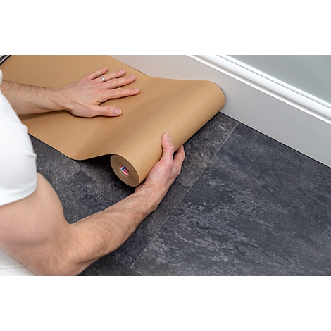 Person is rolling out a paper sheet on a dark floor to protect it from work. White wall in the background.