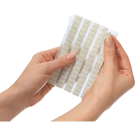 Hands holding a package of small, square adhesive pads in light gray. One pad is being removed.