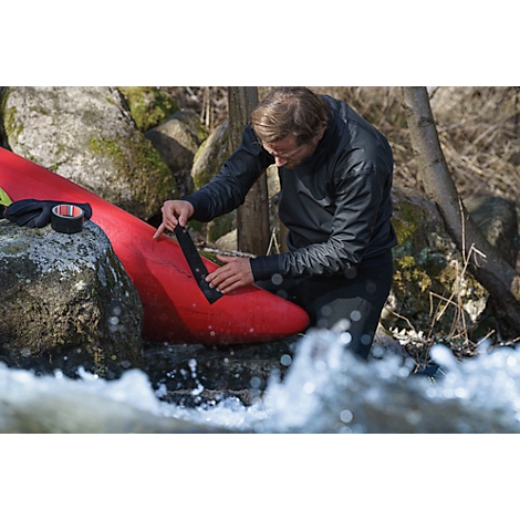 Man taping red kayak. He stands in water in front of rocks and a tree. Tape is ready.
